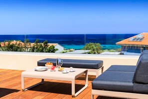 A balcony with a table set for breakfast, a cushioned seating area, and a view of the sea.