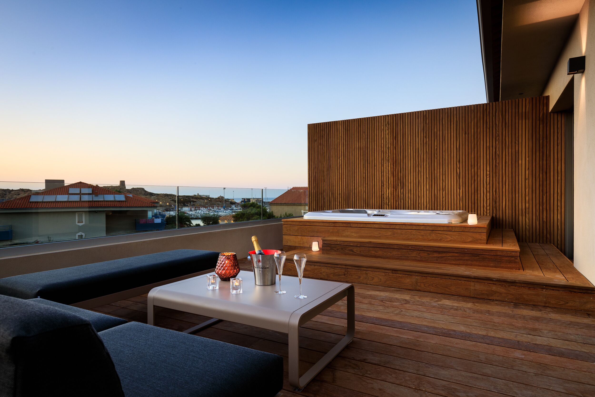 A rooftop terrace with a wooden deck, a seating area with cushions, a table with glasses and a bottle, and a hot tub.
