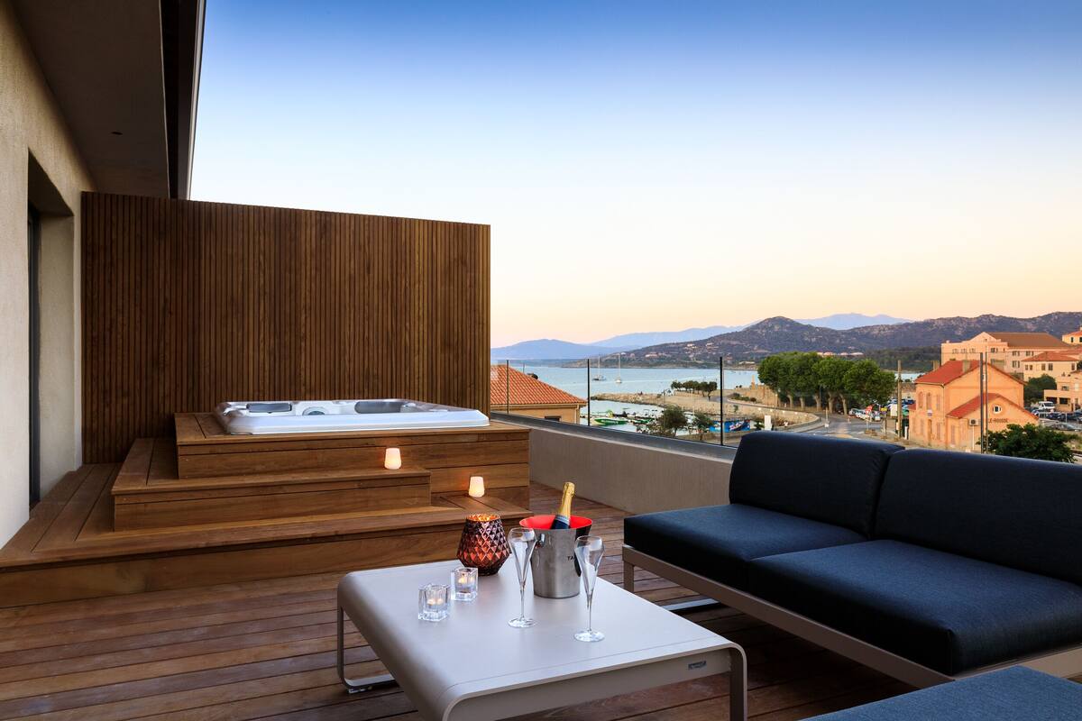 A balcony with a wooden deck, a sofa, a table with glasses and a bottle, and a hot tub.