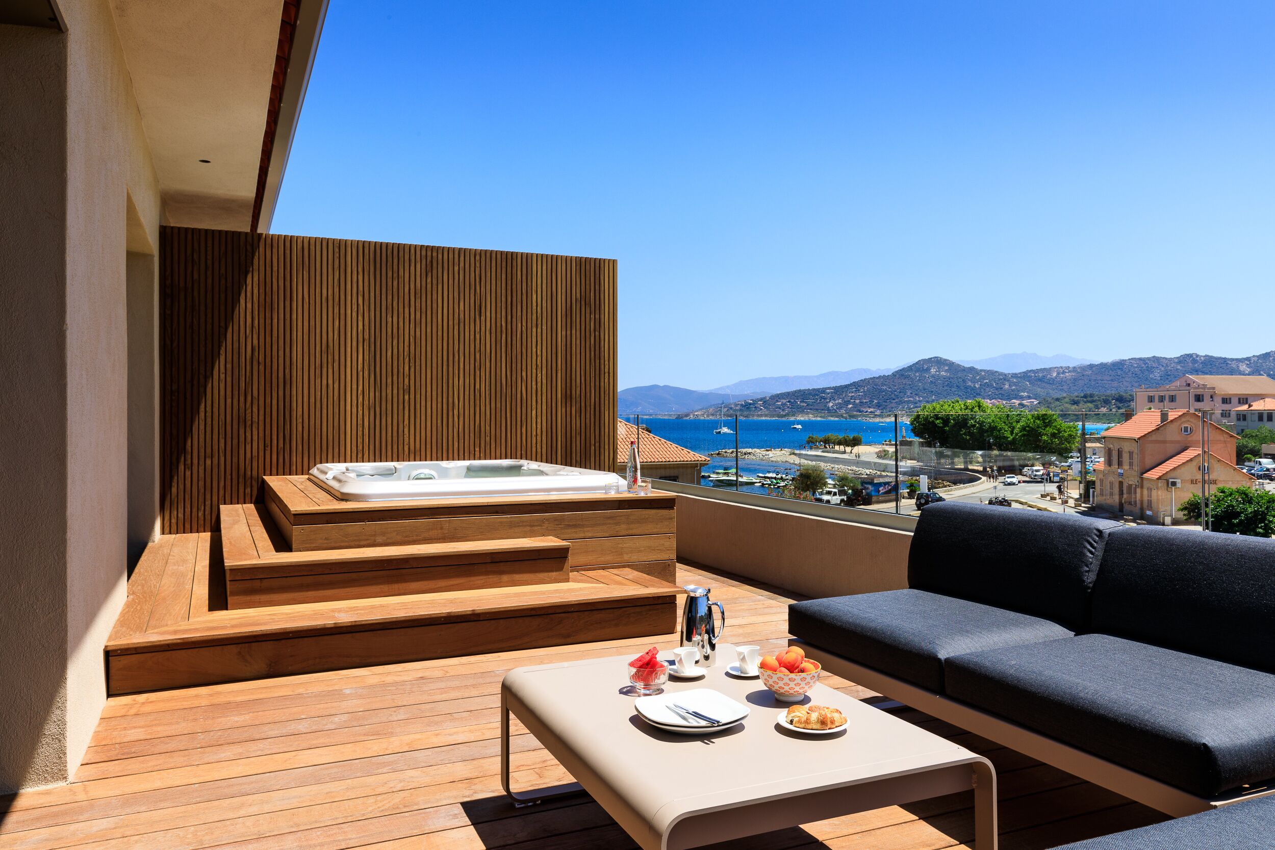 A balcony with a wooden floor, a sofa, a table with breakfast items, and a hot tub.