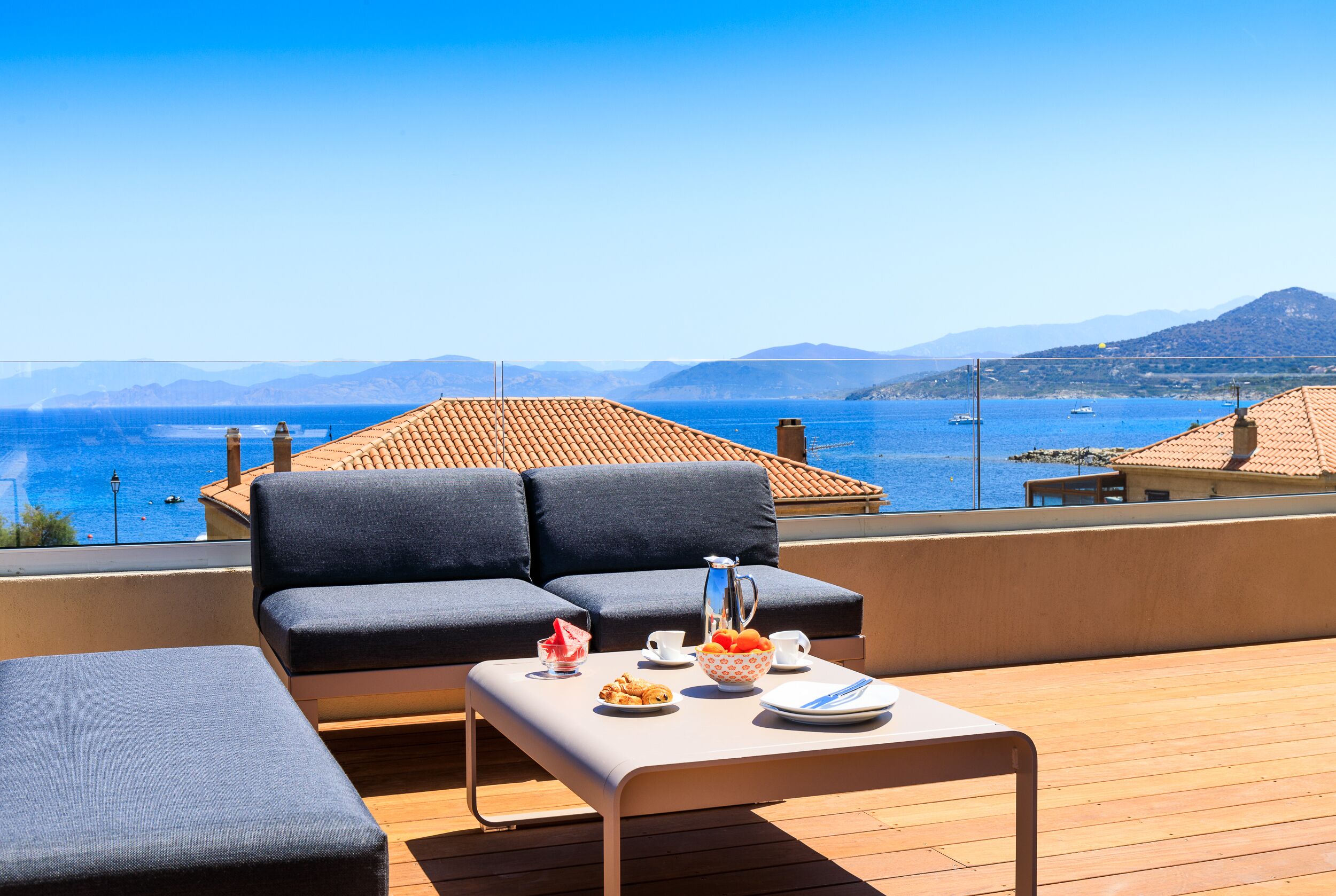 Une terrasse avec un canapé, une table dressée pour le petit-déjeuner et une vue sur la mer et les montagnes.