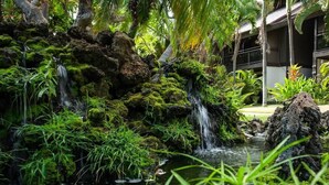 A lush garden with a small waterfall, dense greenery, and a building in the background. - Beach & Poolside Bliss - Family-Friendly Maui Stay (Kihei)