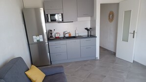 Een moderne keuken met grijze kastjes, een roestvrijstalen koelkast, een magnetron en een koffiezetapparaat. Er staat een grijze bank met een geel kussen en een klein houten tafeltje met een glazen pot.