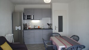 Een compacte keuken met grijze kastjes, een magnetron en een spoelbak. Er is een eettafel met stoelen en een hangende lamp.