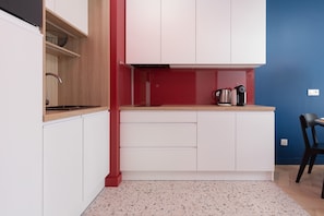 A compact kitchen with white cabinets, a red accent wall, a stainless steel kettle, and a black chair. - Stylish Apartment with Balcony | Parking | 500 m to the Beach (Kołobrzeg)