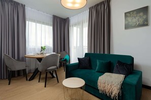 Un salon moderne avec un canapé vert, des fauteuils gris, une table ronde en bois et une petite table d’appoint ronde.