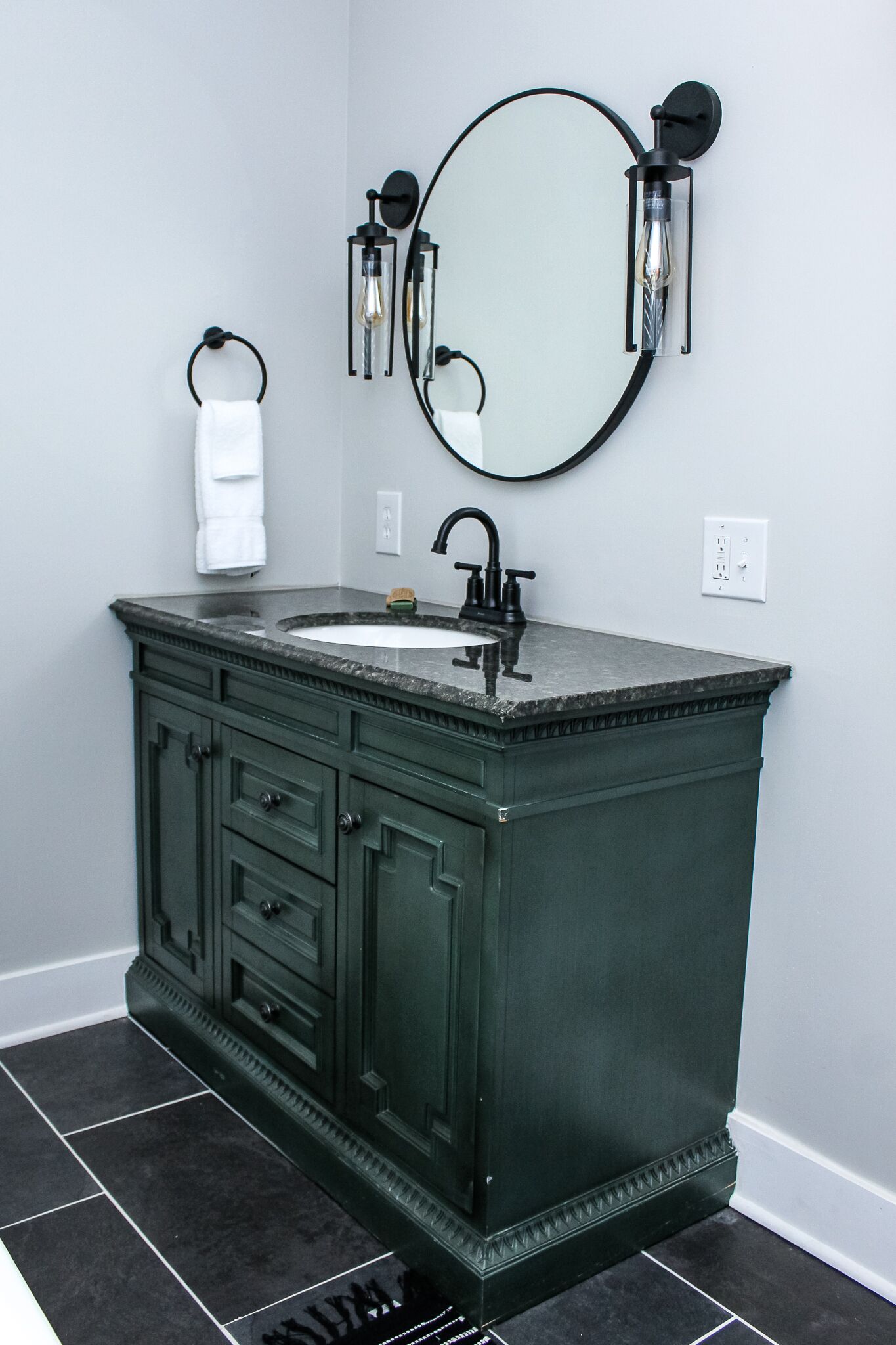 A bathroom with a dark green vanity, a round mirror, and a white sink.