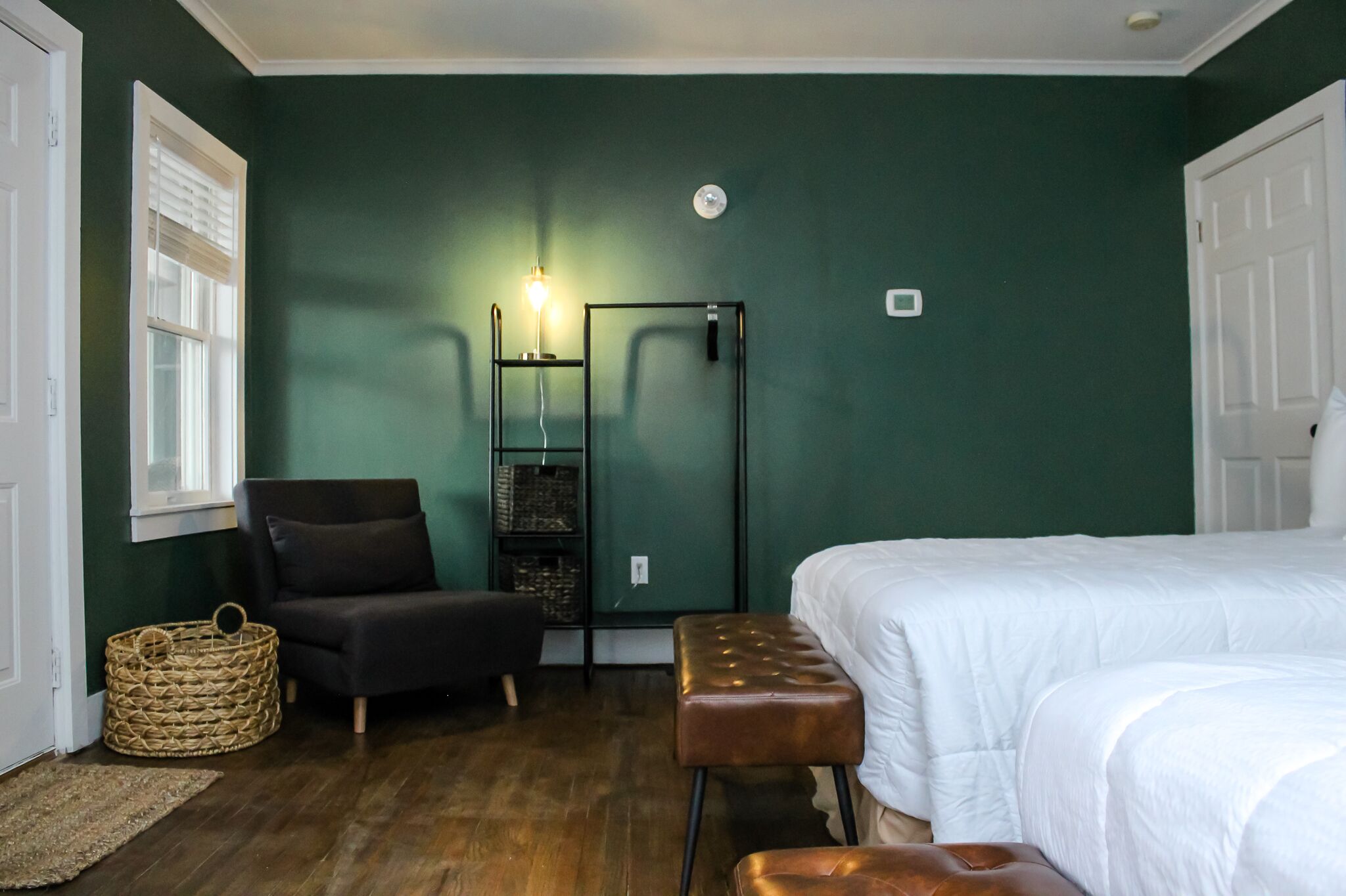 A room with a bed, a chair, a bench, a small table, and a wall-mounted shelf with baskets.