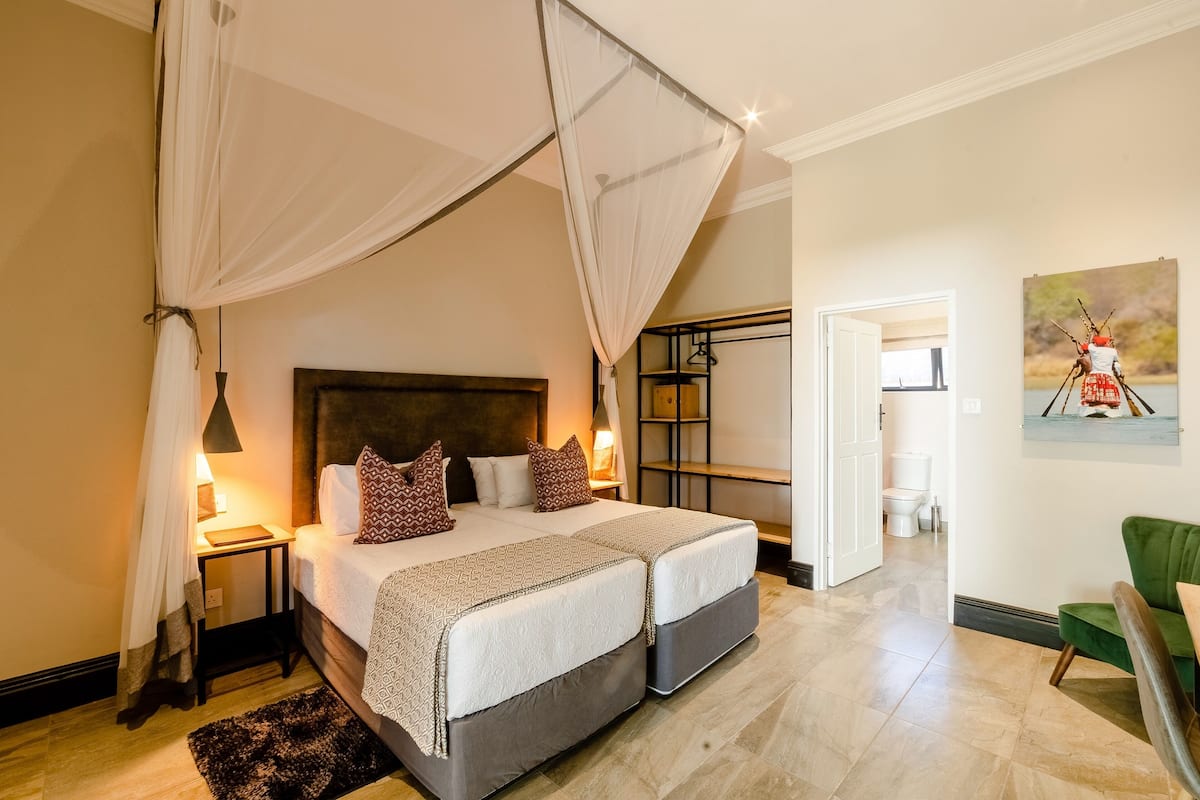 A double bed with a canopy, bedside tables with lamps, a green armchair, and a painting on the wall.