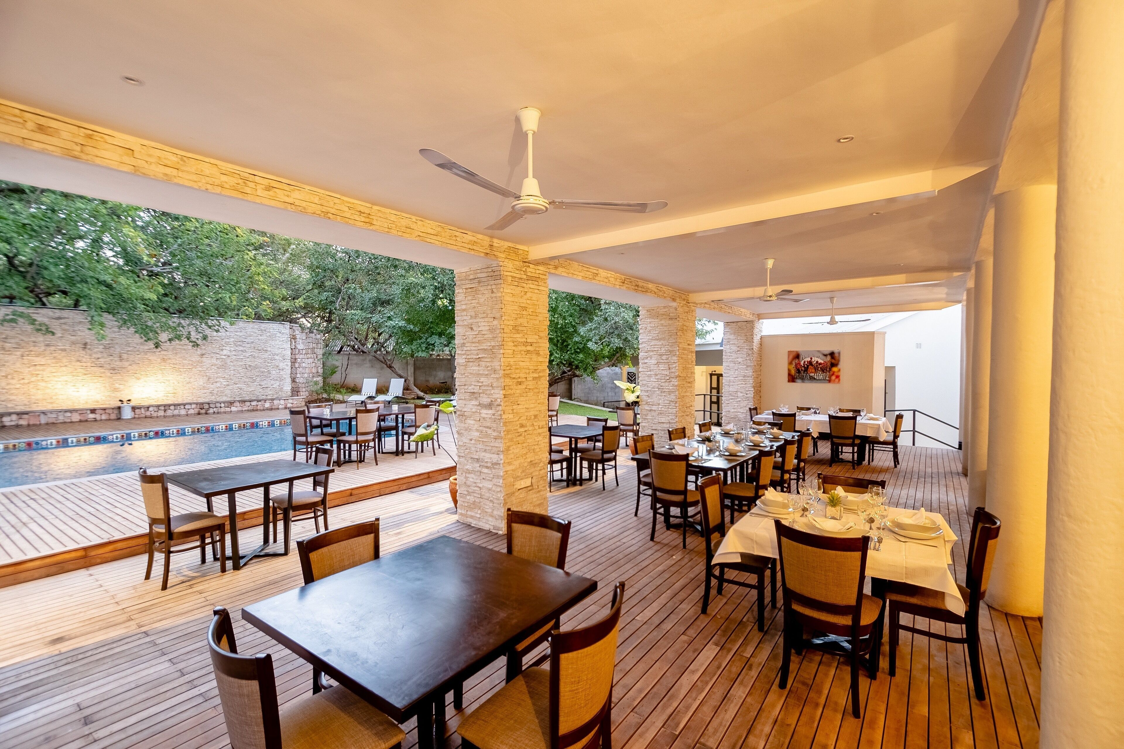 Un área de comedor al aire libre con mesas y sillas de madera, piscina y un muro de ladrillo con plantas.