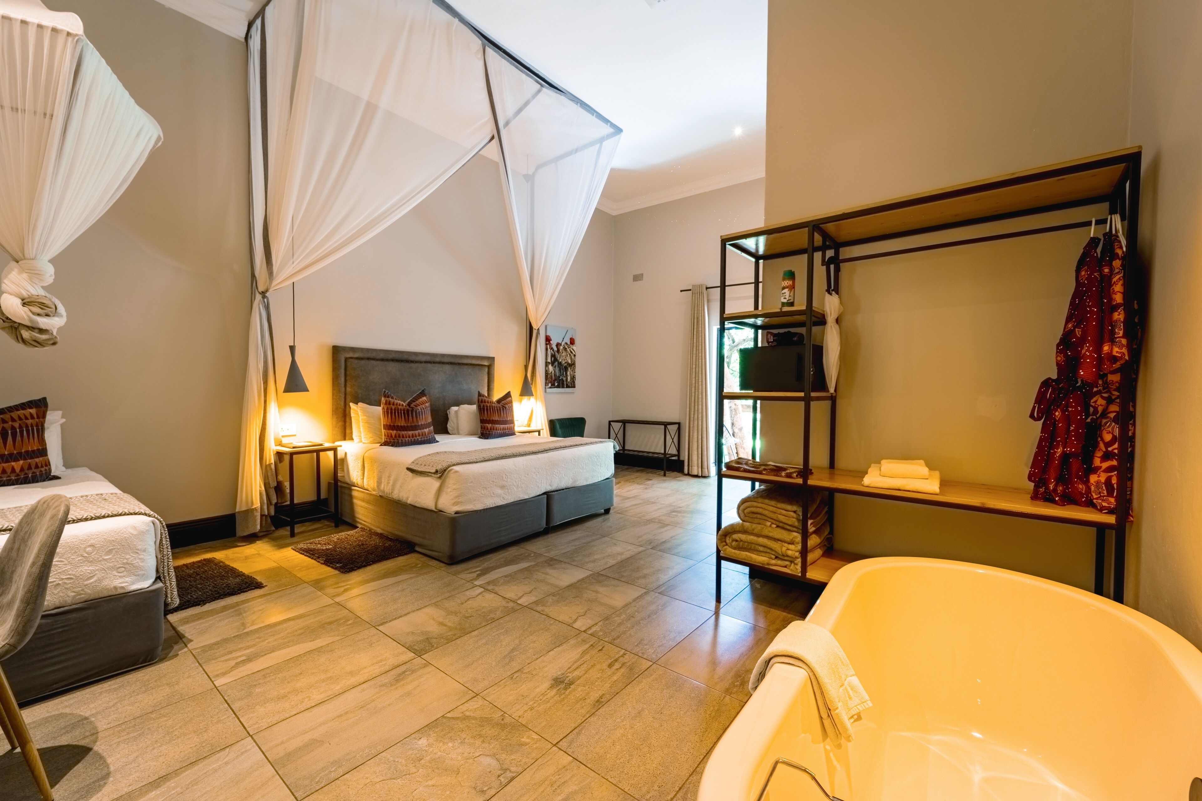 A modern hotel room with a large bed, a sitting area with a chair, a bathtub, and a wardrobe with hanging clothes.