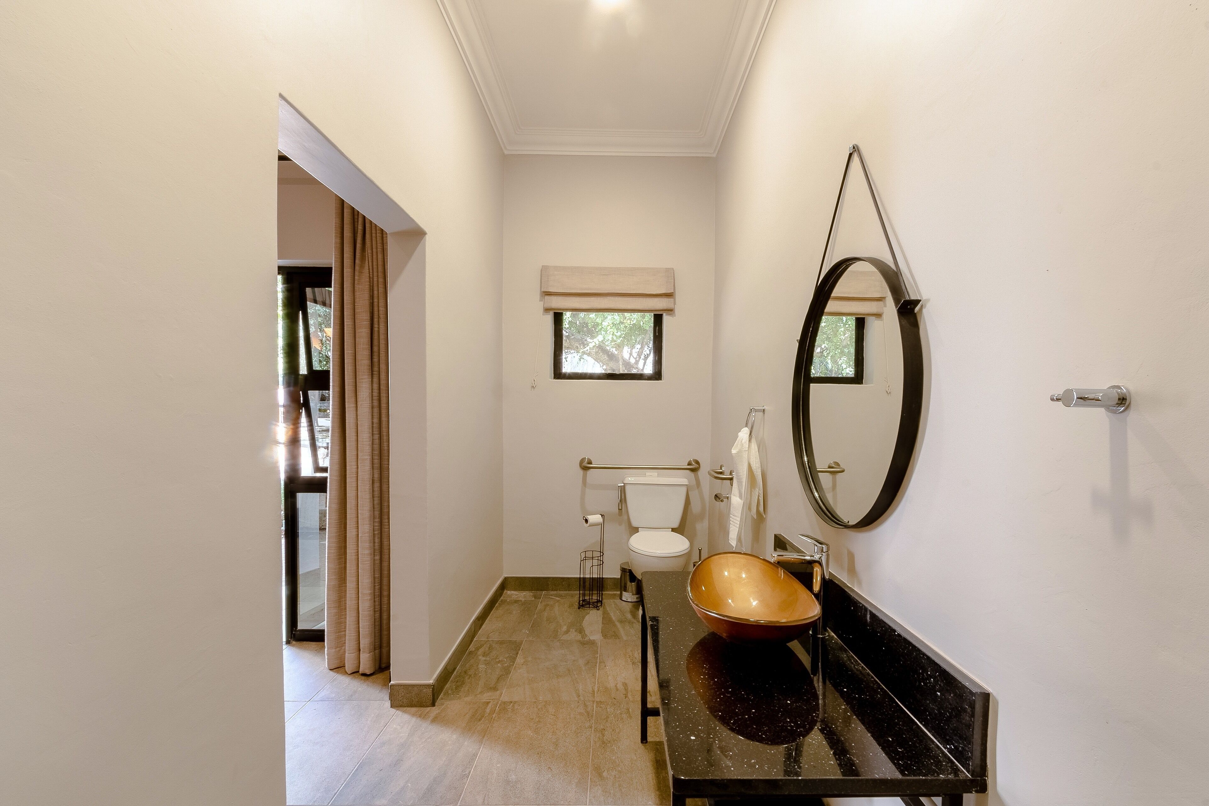 A bathroom with a black countertop, a round mirror, a toilet, and a window with a beige blind.