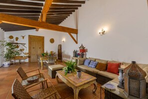 Ein rustikales Interieur mit freiliegenden Holzbalken, einer Essecke mit Holztisch und -stühlen, einer Couch mit Kissen sowie einer Wand mit Wandleuchten.
