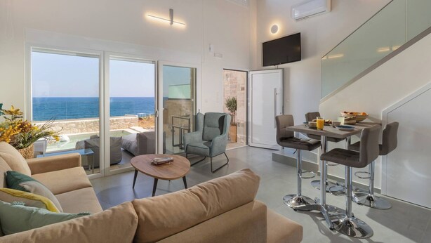 A spacious living area with a large sliding glass door leading to a balcony overlooking the sea, a comfortable seating area, a dining space with bar stools, and a flat-screen TV mounted on the wall.