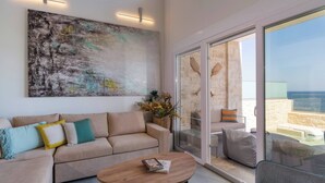 A modern living room with a sofa, a coffee table, and a large abstract painting. There is a sliding glass door leading to an outdoor area with a stone wall and a view of the sea.