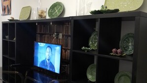 Een kamer met een televisie op een standaard, omringd door planken met decoratieve borden en een boekenkast.