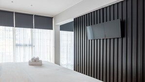 Una habitación de hotel moderna con una cama grande, un televisor de pantalla plana montado en una pared de madera oscura y ventanas amplias con cortinas de gasa.