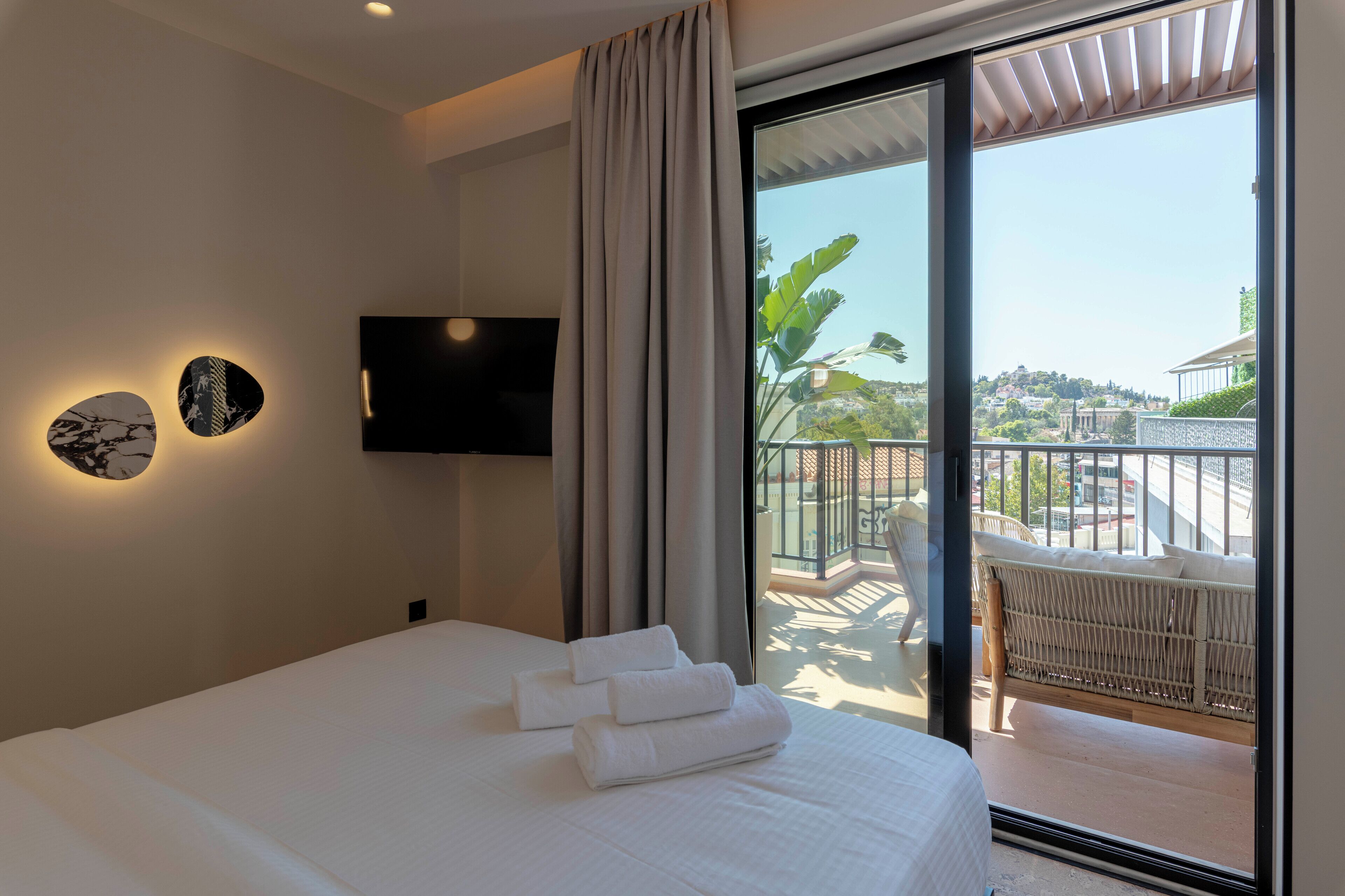 A hotel room with a bed, a flat-screen TV mounted on the wall, and a balcony with a wicker chair.