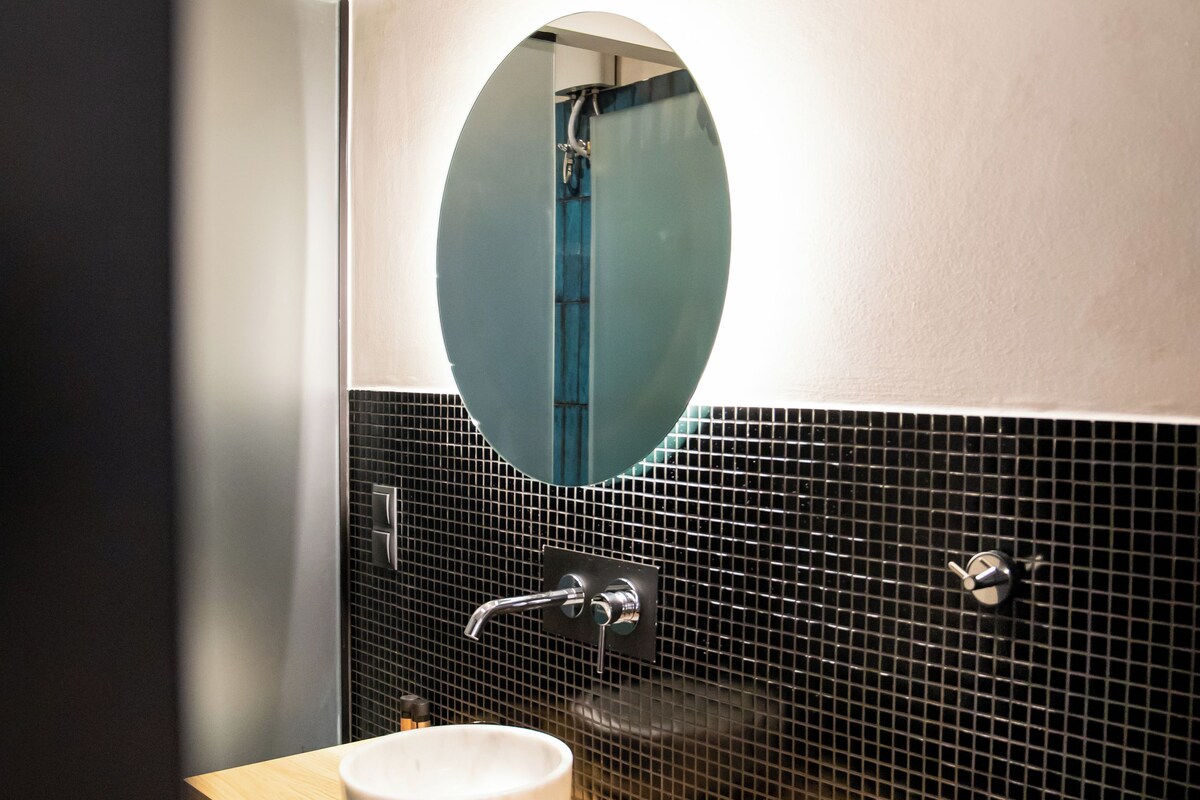 A modern bathroom with a wooden vanity, a round mirror, and black tiled walls.