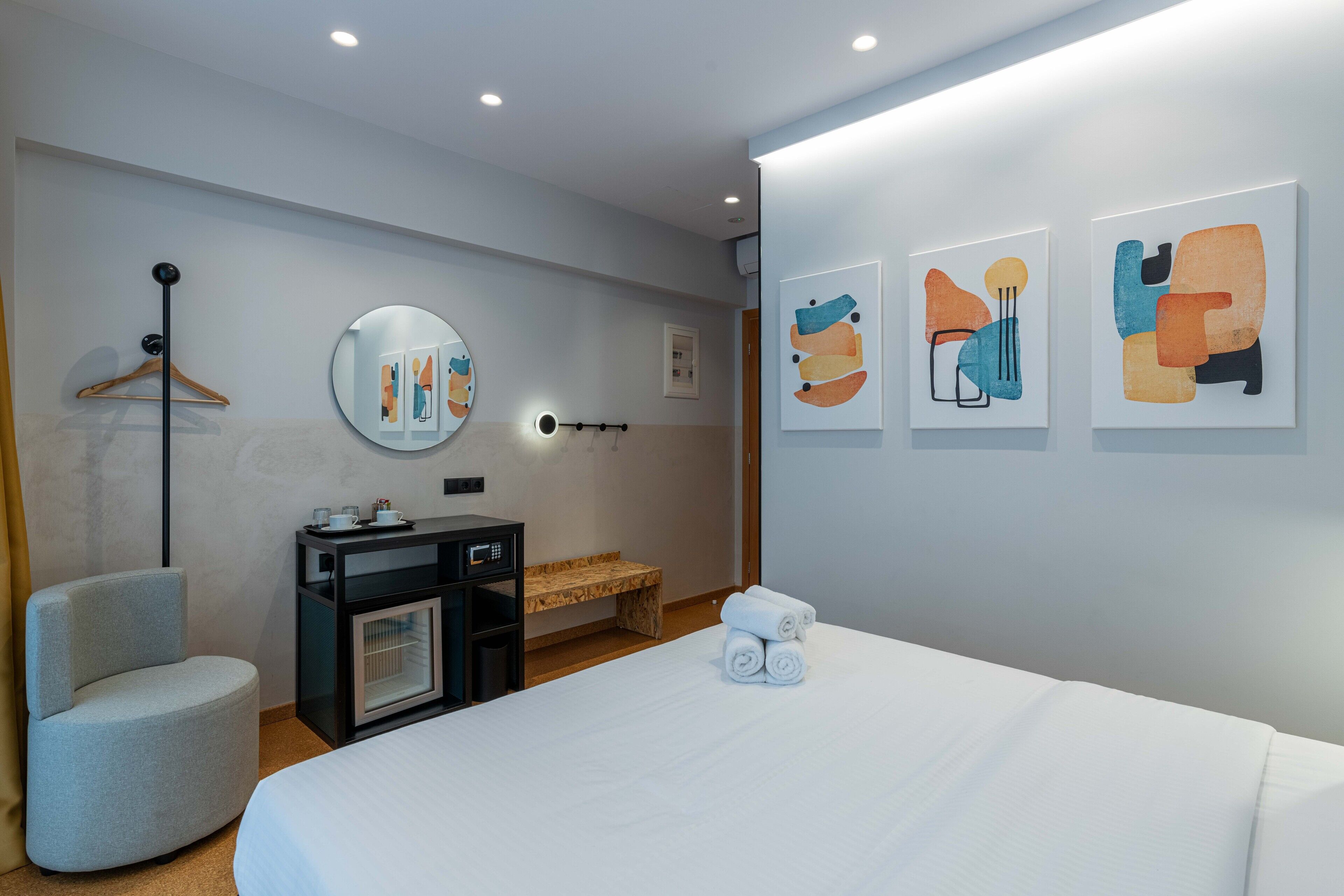Et moderne hotelværelse med seng, natbord, stol, skrivebord, spejl og abstrakt kunst på væggen.