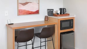 A kitchenette with a microwave, coffee maker, and refrigerator. There are two bar stools and a painting on the wall.