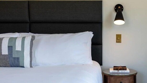 A neatly made bed with white linens and a striped pillow, a dark upholstered headboard, a bedside table with a lamp, and a small decorative item on the table.