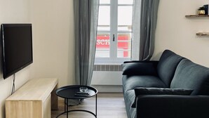 Ein modernes Wohnzimmer mit einem schwarzen Sofa, einer Holzbank, einem kleinen runden Tisch, einem Flachbildfernseher und einem Fenster mit grauen Vorhängen.