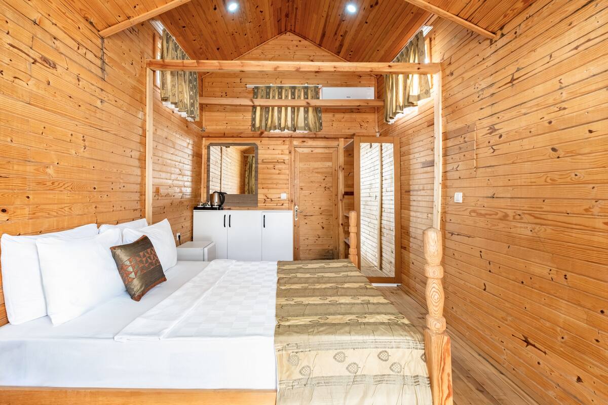 A wooden cabin interior with a bed, a small kitchenette, and a window.