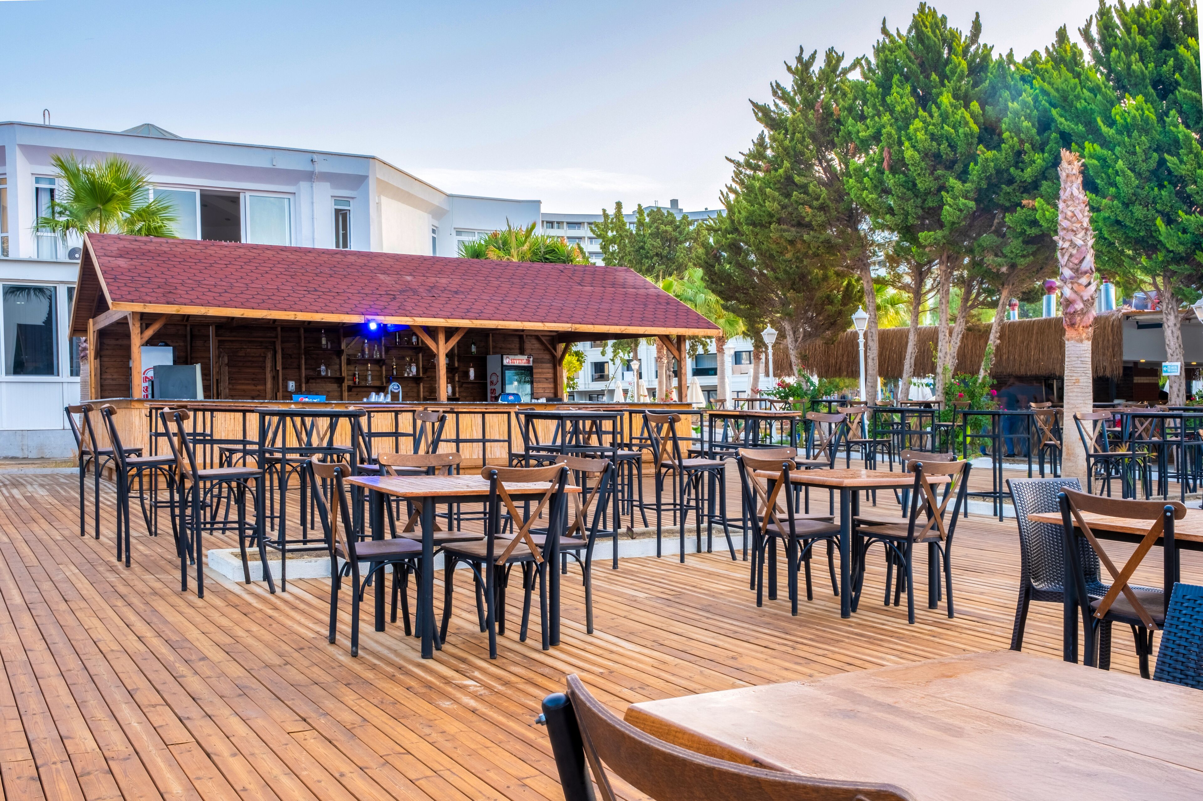 Un área de comedor al aire libre con mesas y sillas de madera, una zona de bar cubierta y vistas a edificios y árboles al fondo.