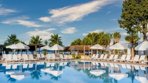 Una piscina azul cristalina con sillas de descanso blancas y sombrillas, rodeada de palmeras y una estructura de techo de paja.