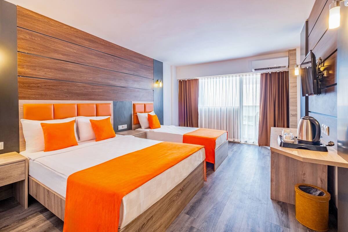 A hotel room with two beds, a wooden headboard, a small desk with a kettle, and a TV mounted on the wall.