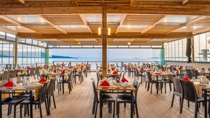 Un amplio comedor al aire libre con mesas y sillas de madera, grandes ventanales con vistas al mar y una estructura de techo de madera.