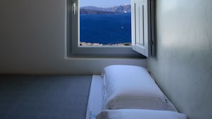 A small bedroom with a single bed, a window with an open shutter, and a view of the sea and mountains. - Acro Blue Luxury Villas (Santorini)