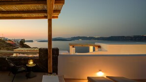 A terrace with a view of a body of water, a stone wall, a wooden post, a chair, a table, and a lamp. - Acro Blue Luxury Villas (Santorini)