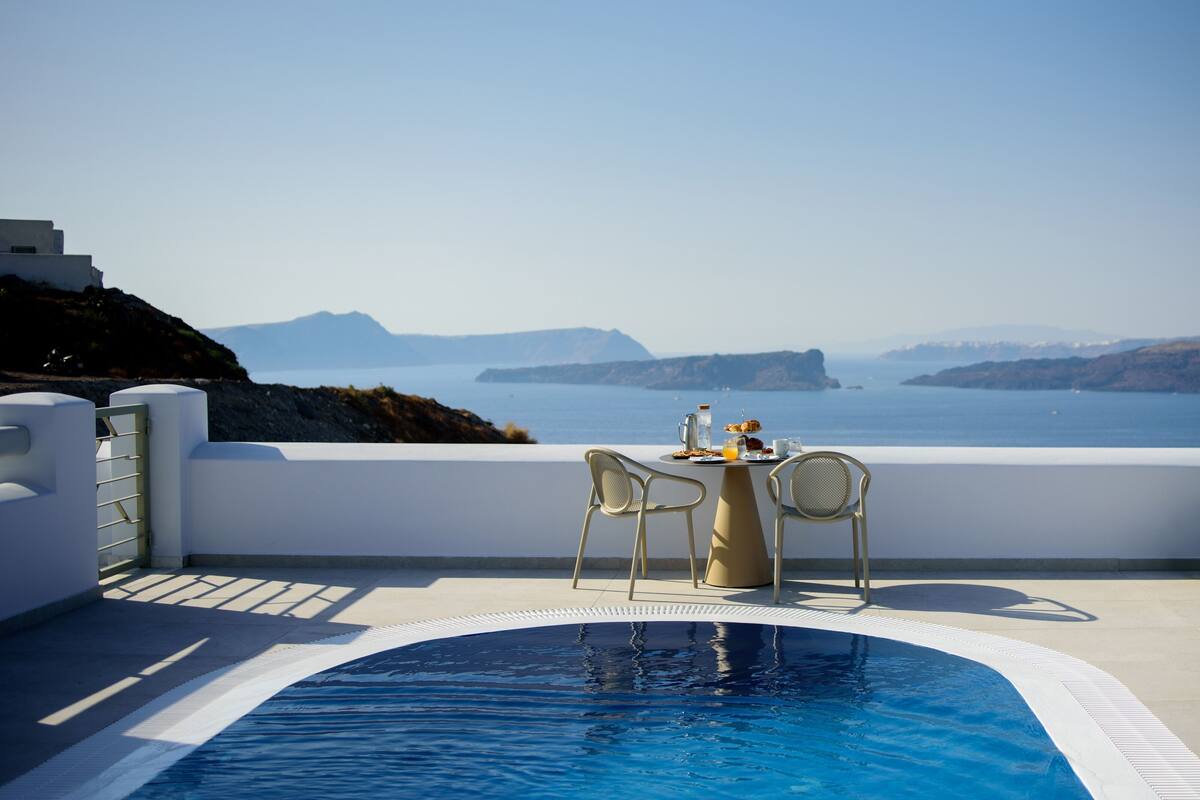 Uma área à beira da piscina com mesa e cadeiras, com vista para um corpo d'água e paisagens distantes.