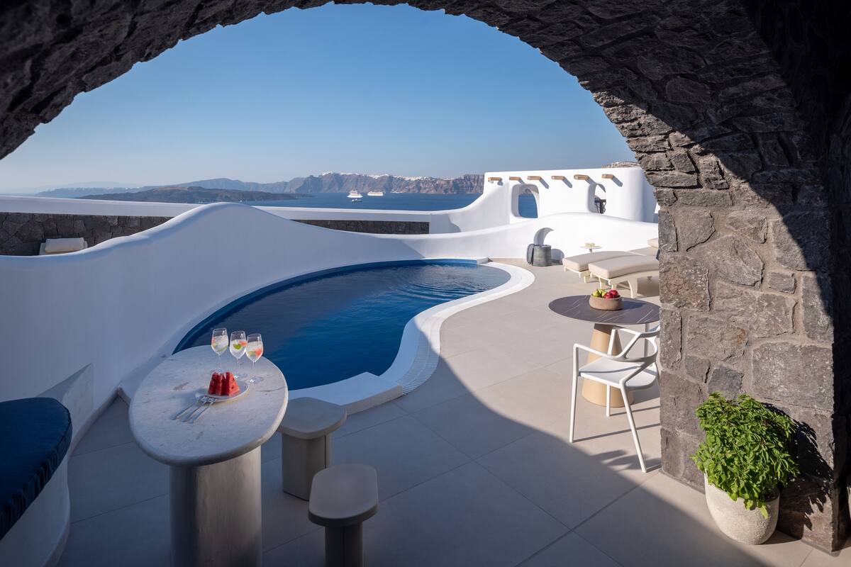 Área da piscina com vista para o mar, paredes de pedra e uma mesa com bebidas.