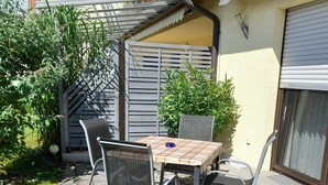 Une terrasse avec une table et des chaises, entourée de verdure et donnant sur un bâtiment doté d’une fenêtre.