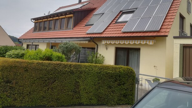 Une maison avec des panneaux solaires sur le toit, un petit balcon et une haie bien taillée dans le jardin.