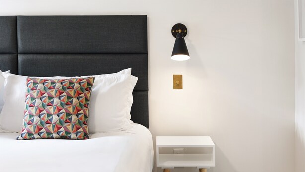 A neatly made bed with white bedding and a colorful geometric pillow, a black upholstered headboard, a white bedside table with a small drawer, and a wall-mounted light fixture.