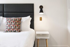 Un lit bien fait, avec une literie blanche et un coussin aux motifs géométriques colorés, une tête de lit noire en tissu, une table de chevet blanche avec un petit tiroir et un luminaire fixé au mur.
