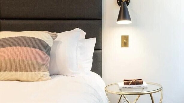 A neatly made bed with white bedding and striped pillows, a round glass side table with a book, and a wall-mounted lamp.