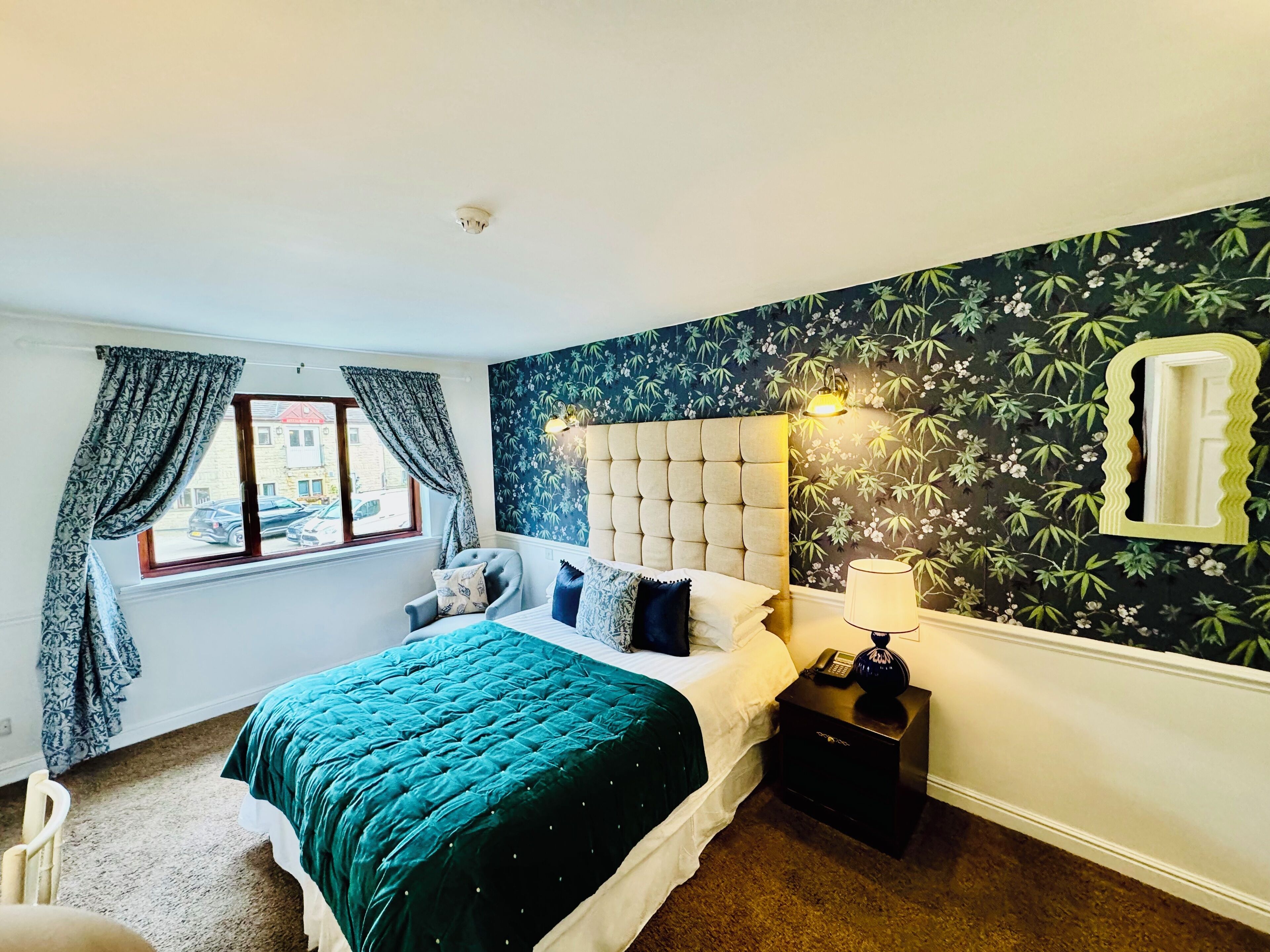 Un dormitorio con una cama, una mesita de noche, una lámpara, una ventana con cortinas y un papel tapiz con motivos florales.