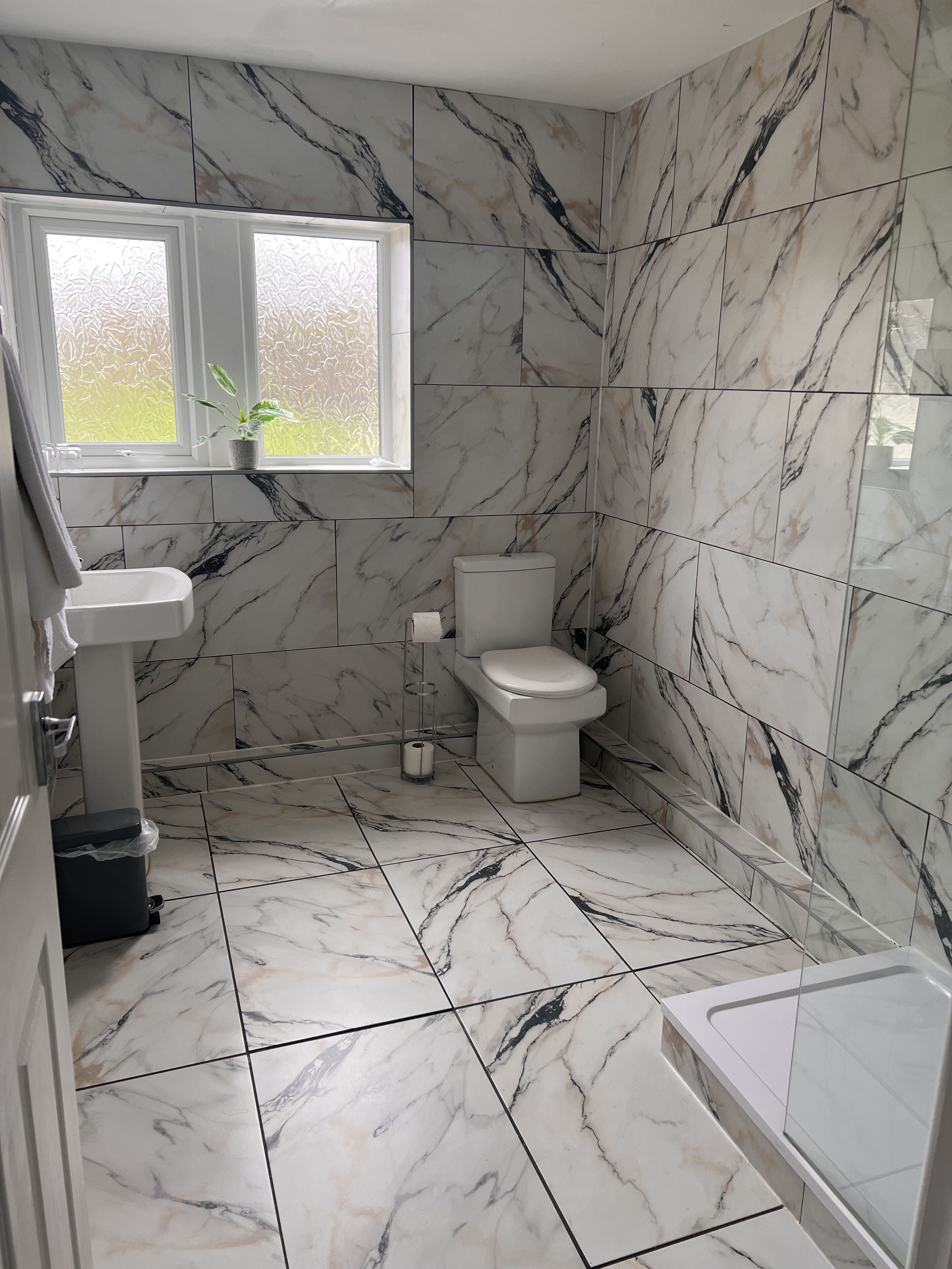 A bathroom with marble tiles, a toilet, a sink, and a window with a view of greenery.