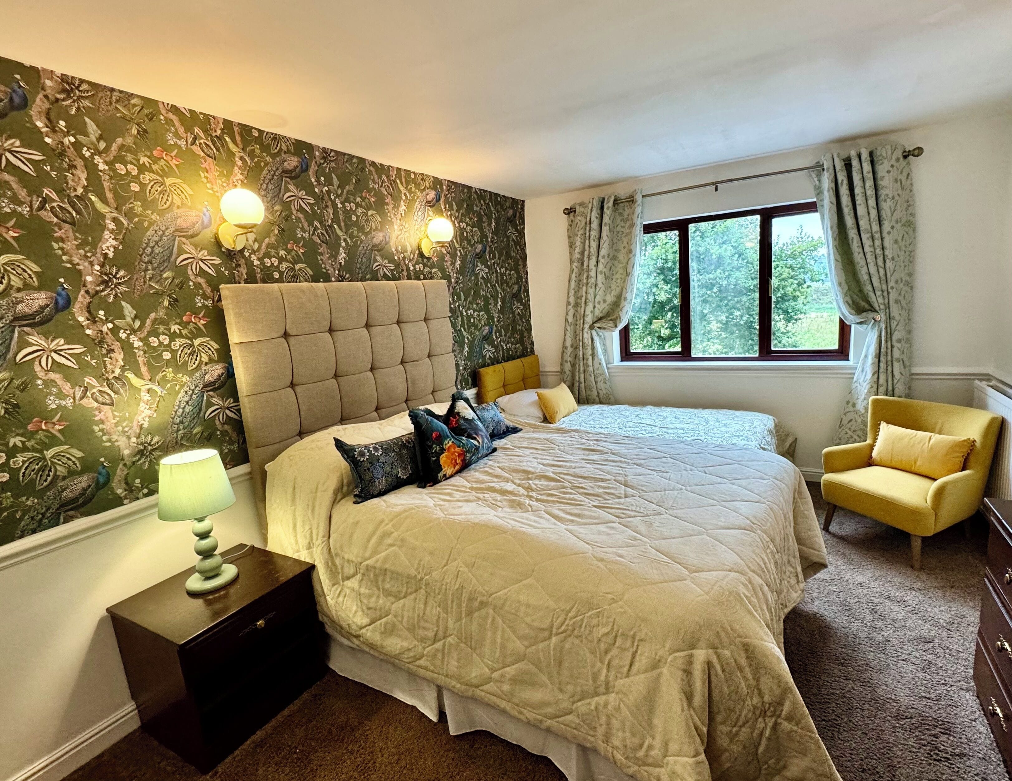 A bedroom with a large bed, a nightstand, a lamp, a window with curtains, and a yellow armchair.