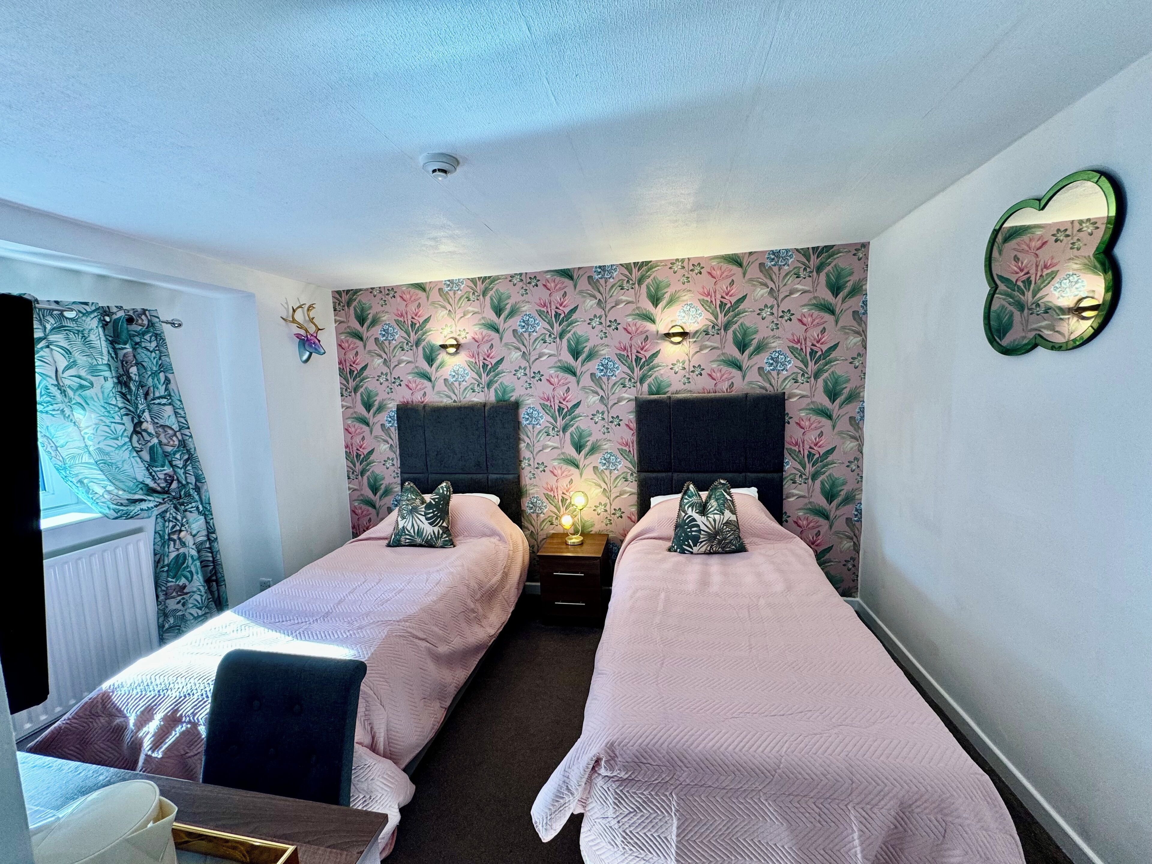 A hotel room with two single beds, a desk, a chair, a window with curtains, floral wallpaper, and a ceiling light.