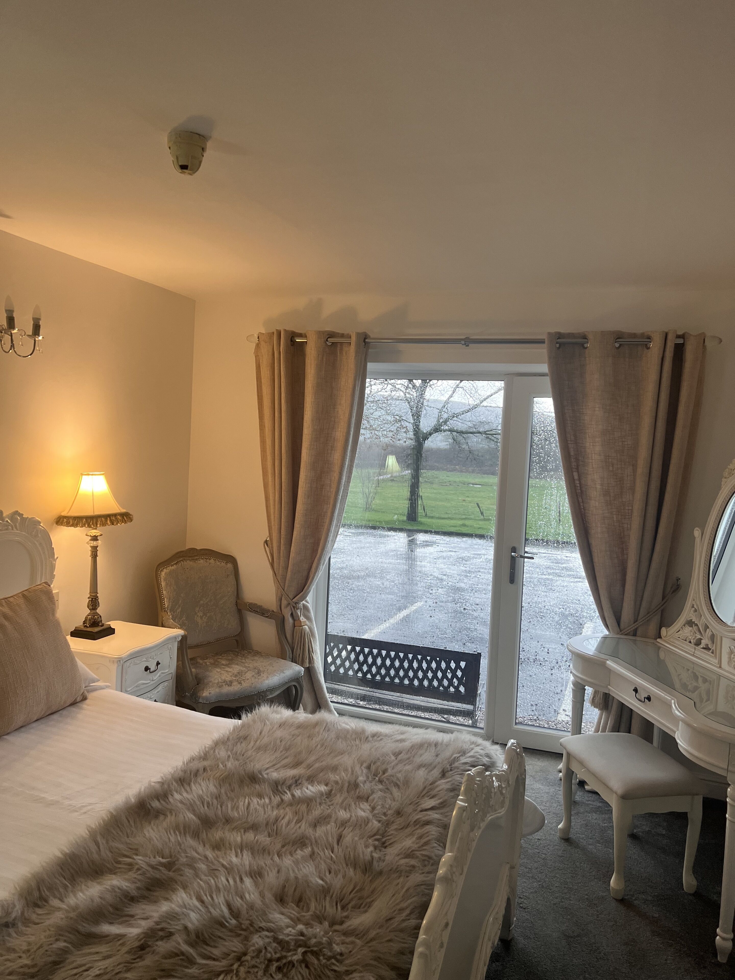 A hotel room with a bed, a desk, a chair, a lamp, and a view of the outdoors.