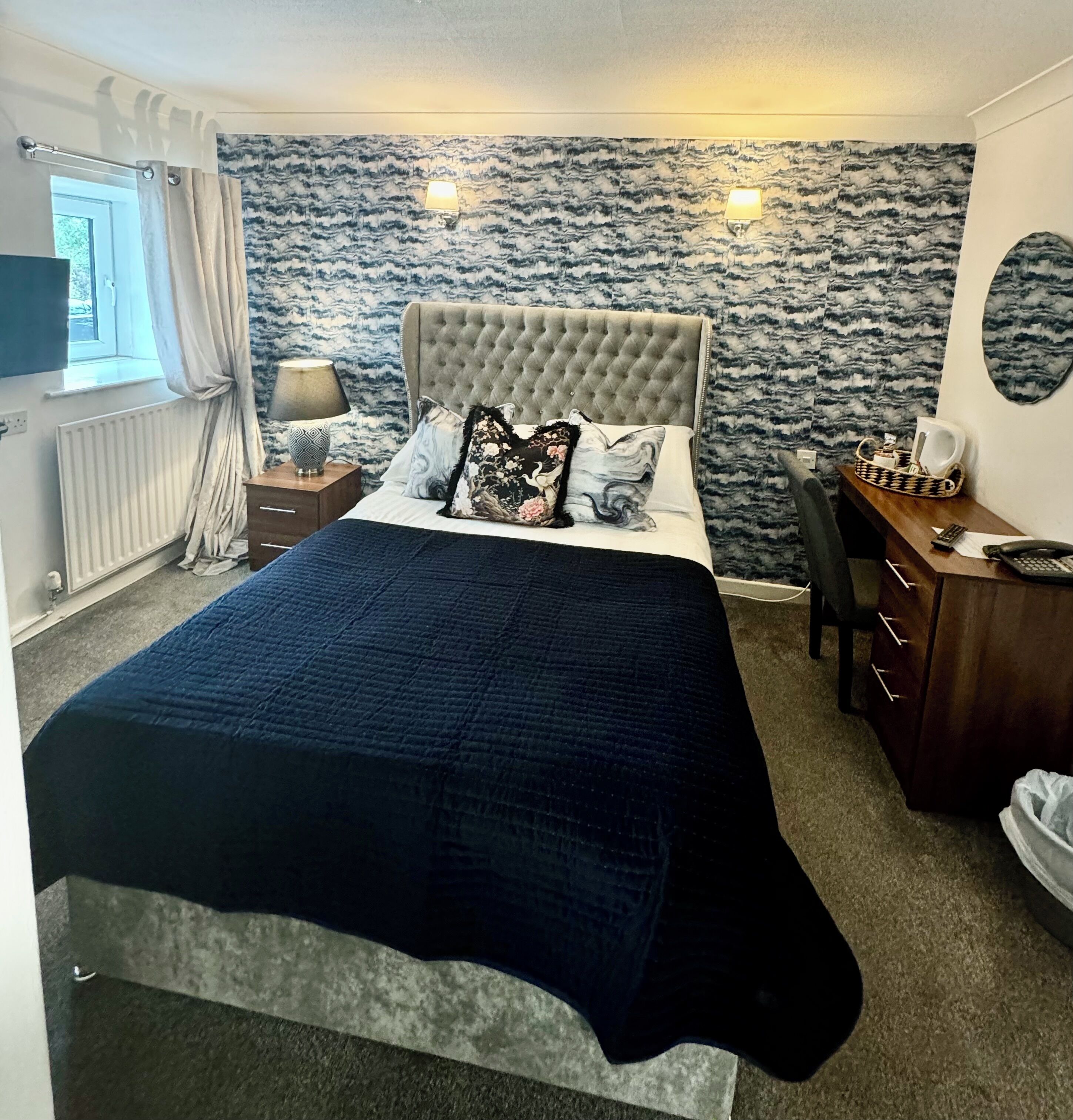 A bedroom with a large bed, a desk, and a chair. There is a window with curtains, a lamp, and a radiator.