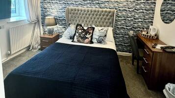 A bedroom with a large bed, a desk, and a chair. There is a window with curtains, a lamp, and a radiator.