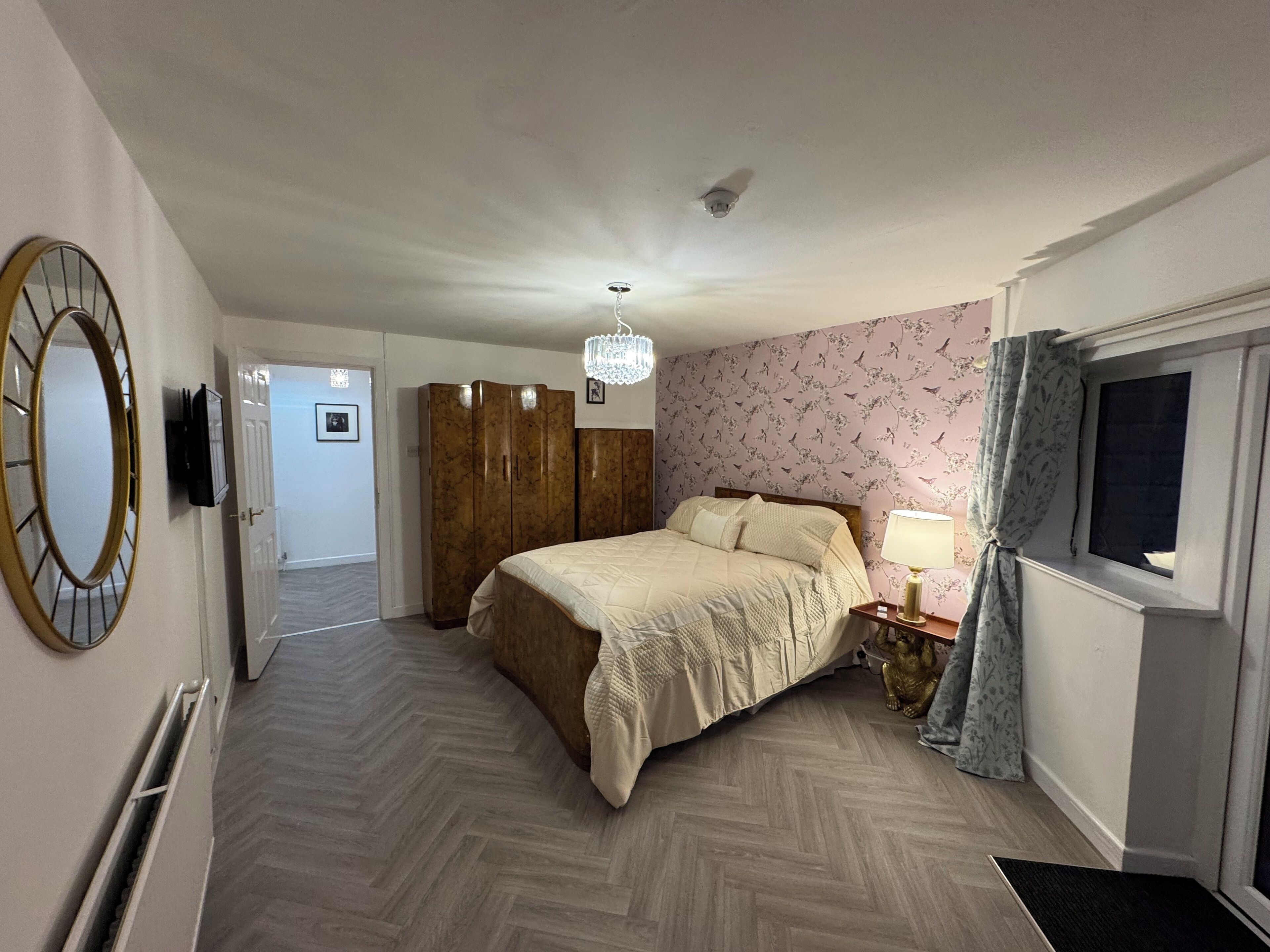 Een slaapkamer met een bed, een kledingkast, een spiegel, een kroonluchter, een raam met gordijnen en een deur naar een andere kamer.