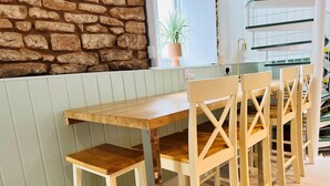 Une table en bois, assortie à des chaises, se trouve dans une pièce dont un mur est en pierre et qui possède une fenêtre ornée d’une plante.